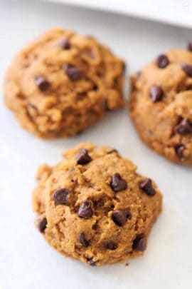 Three chocolate chip cookies on a white surface. They have a light brown color with visible chocolate chips.