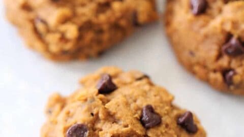 Three chocolate chip cookies on a white surface. They have a light brown color with visible chocolate chips.