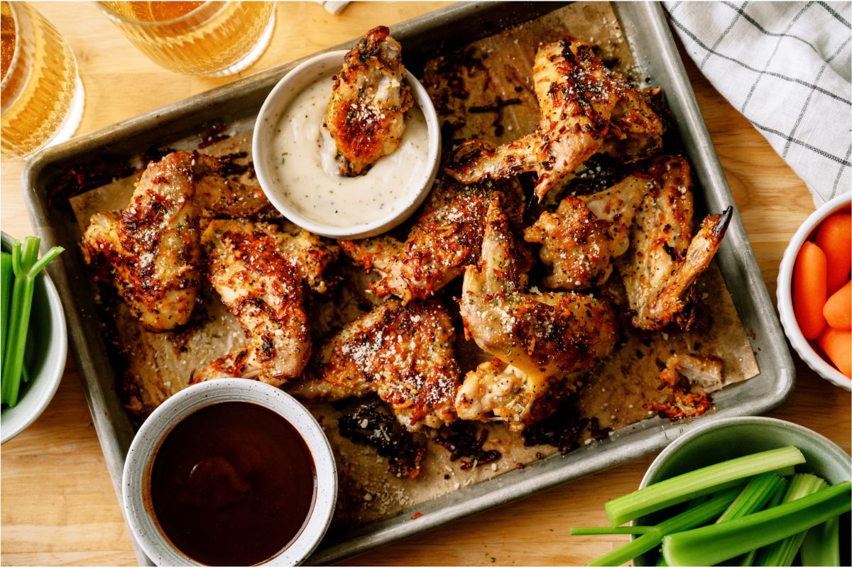 Top view of a sheet pan of Air Fryer Garlic Parmesan Chicken Wings with 2 cups of dipping sauces on the pan. One of the dipping sauces has a wing in it. Carrots and Celery Sticks in bowls off to the side.