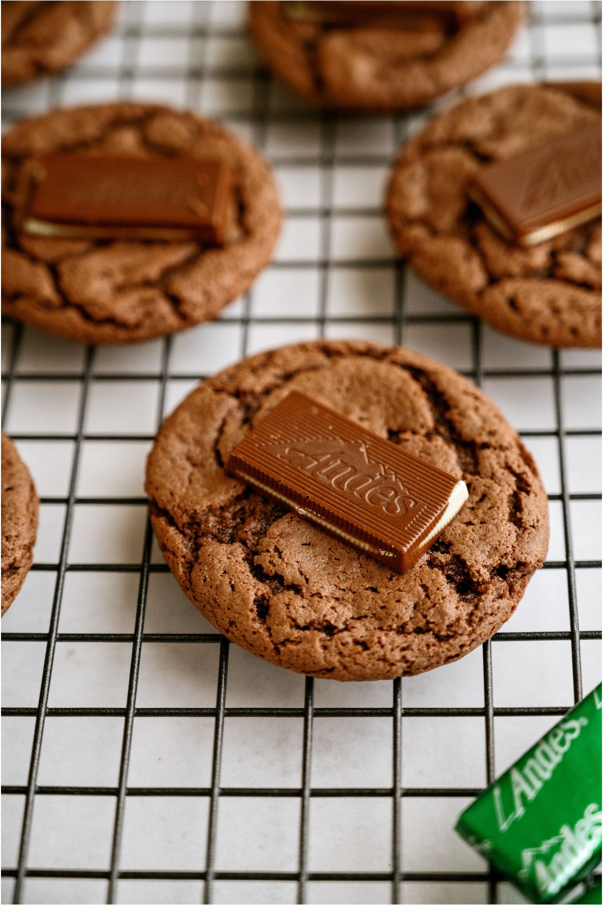 Chocolate cookies on a cooling rack with an unwrapped andes mint on top of it. Other cookies with mints on top in the background.