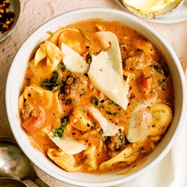 A bowl of creamy tomato pasta soup with tortellini, ground meat, spinach, and slices of parmesan cheese, served with a side of butter.