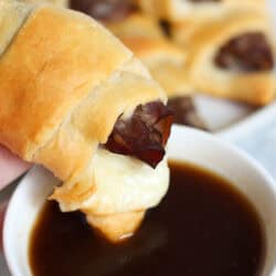 A hand holding a cooked french dip crescent over a small bowl of Au Jus with a bite missing from the crescent.