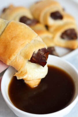 A hand holding a cooked french dip crescent over a small bowl of Au Jus with a bite missing from the crescent.