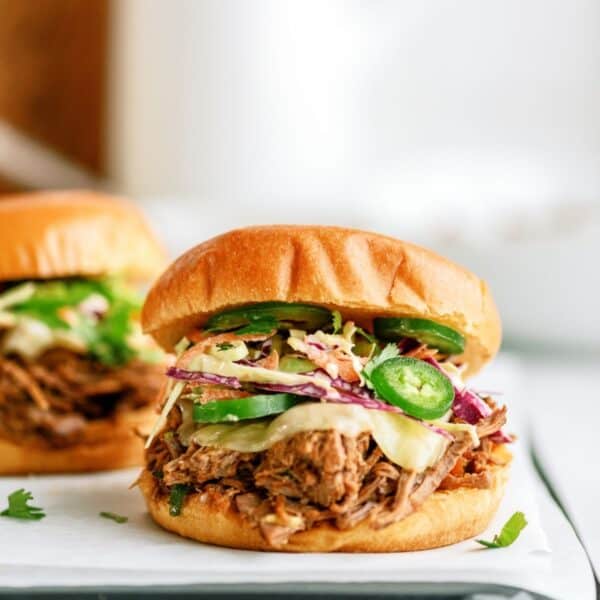 A pulled pork sandwich with coleslaw, sliced jalapeños, and a white sauce in a bun is placed on a white surface.