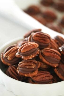 A white bowl is filled with pecan halves sandwiching chocolate, with more of the same in the background.