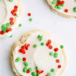 Sugar cookie with white frosting and green and red sprinkles. 2 other sugar cookies with sprinkles off to the side.