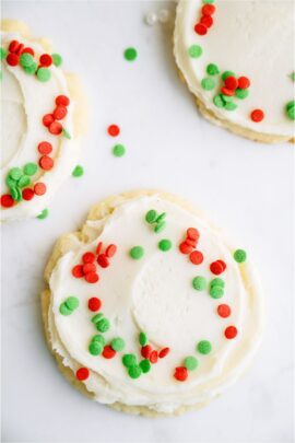 Sugar cookie with white frosting and green and red sprinkles. 2 other sugar cookies with sprinkles off to the side.