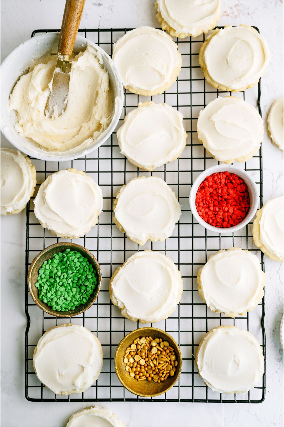 Super Soft Sugar Cookies frosted on a cooling rack with a bowl of frosting and 3 little bowls of different colored sprinkles.