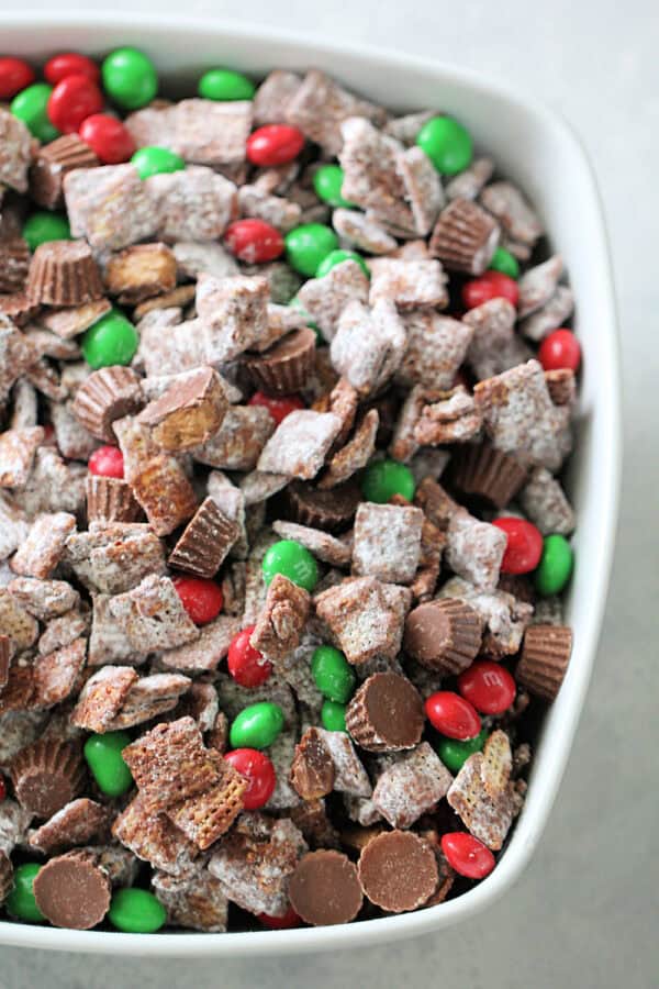 A white bowl filled with chocolate muddy buddies, mini peanut butter cups, and red and green candy-coated chocolates.