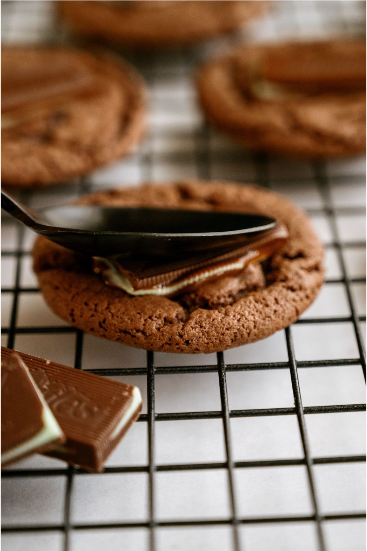 A spoon spreading the melted mint on top of the chocolate cookie on a cooling rack.