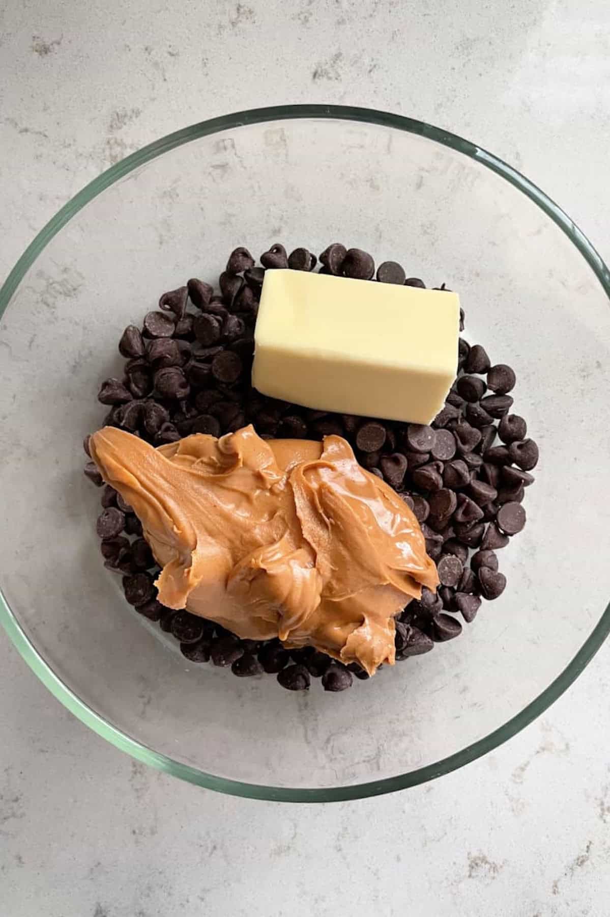 A glass bowl containing chocolate chips, a stick of butter, and a scoop of peanut butter.