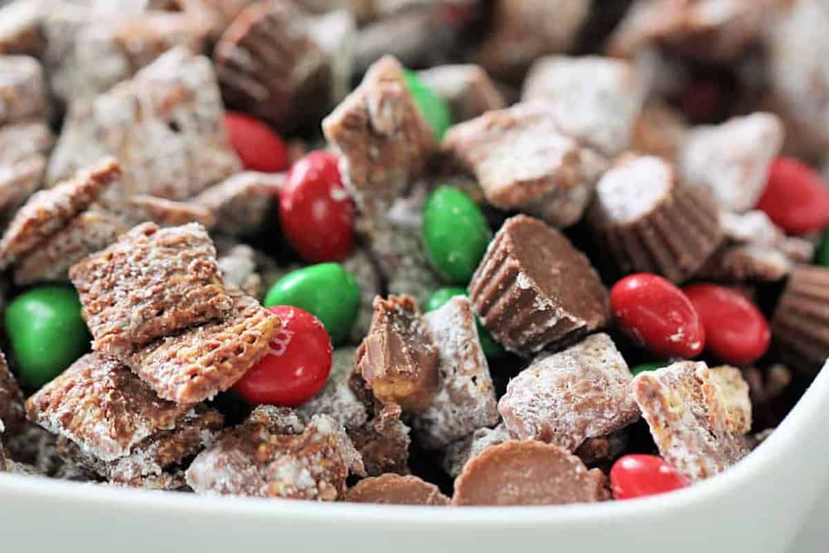 A bowl of snack mix with chocolate-covered cereal squares, red and green candy-coated chocolates, and mini peanut butter cups.