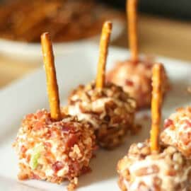 Small cheese balls coated with chopped nuts and bacon, each with a pretzel stick in the center, arranged on a white plate.