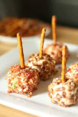 Small cheese balls coated with chopped nuts and bacon, each with a pretzel stick in the center, arranged on a white plate.