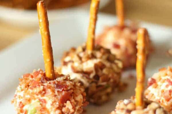 Small cheese balls coated with chopped nuts and bacon, each with a pretzel stick in the center, arranged on a white plate.