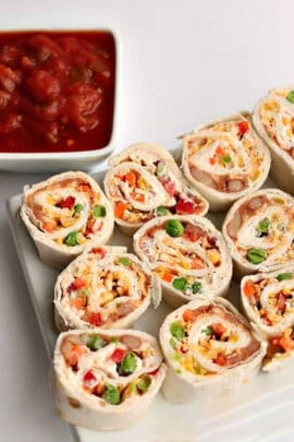 A plate of vegetable and cheese wrapped pinwheels next to a square dish of tomato salsa.