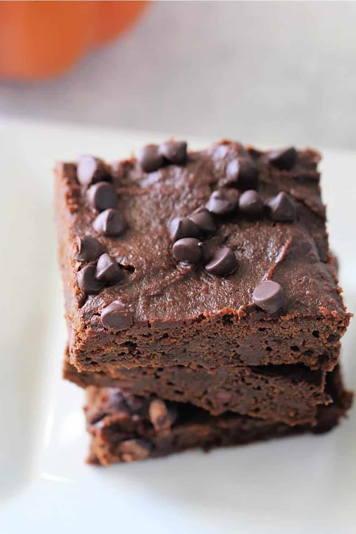 Two chocolate pumpkin brownies are stacked on a white plate, topped with chocolate chips.