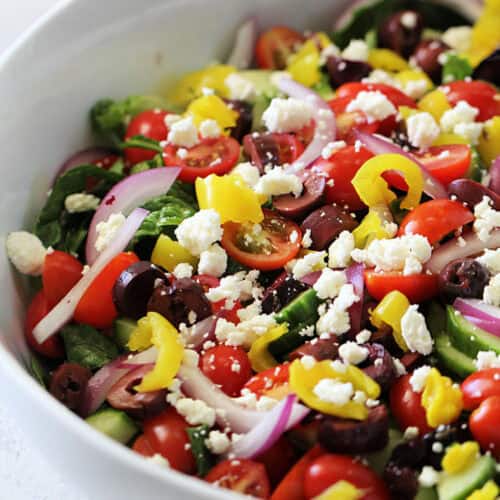 A close-up of a salad with sliced red onions, cherry tomatoes, black olives, yellow bell peppers, cucumbers, and crumbled feta cheese in a white bowl.