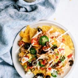 A plate of nachos topped with shredded chicken, sliced jalapeños, black olives, green onions, cherry tomatoes, and drizzled with white sauce, placed on a blue cloth.