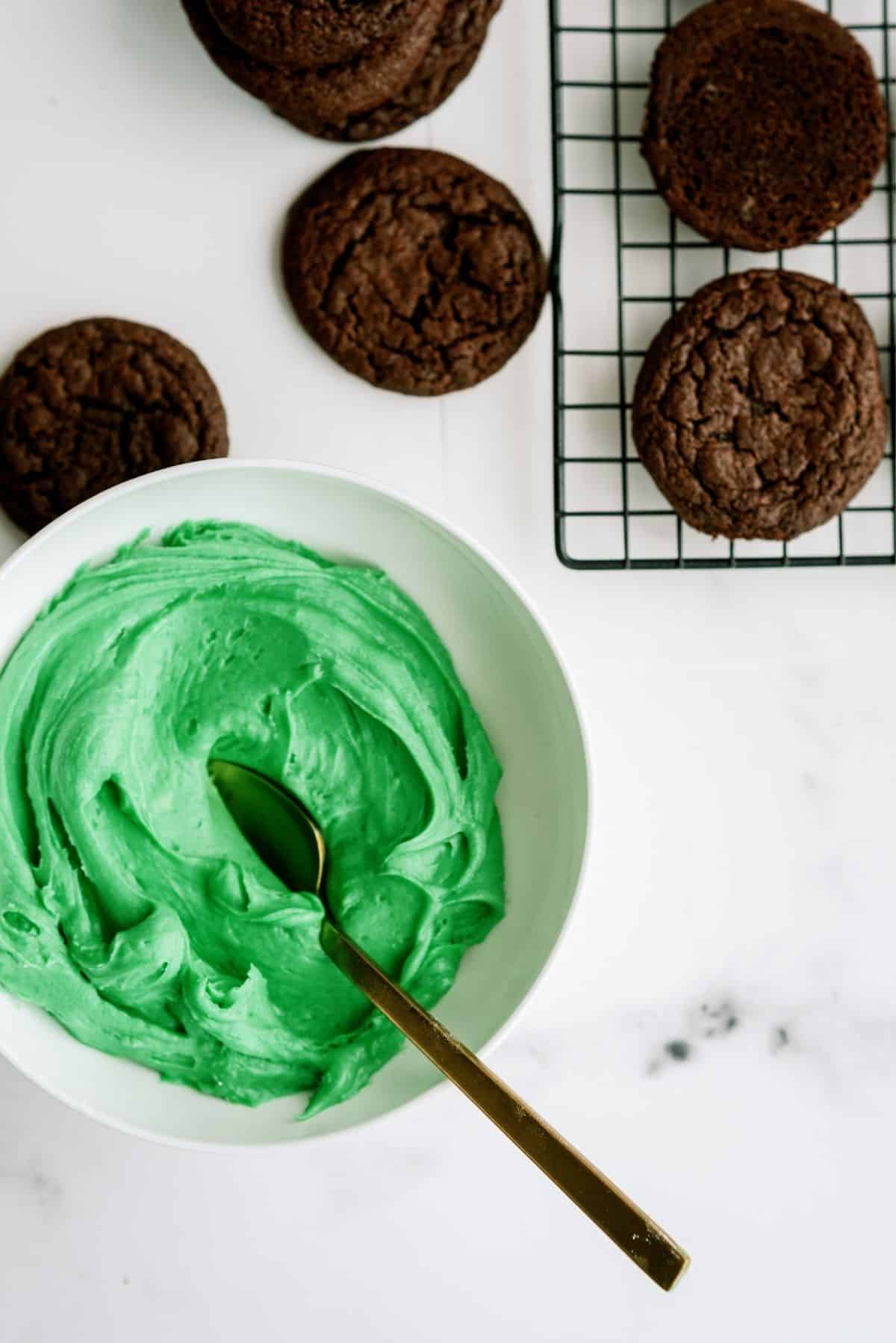 Chocolate cookies on a cooling rack and a few on the counter with a glass bowl full of green icing with a spoon in it.