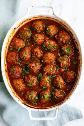 White casserole dish filled with meatballs in tomato sauce, topped with chopped parsley.