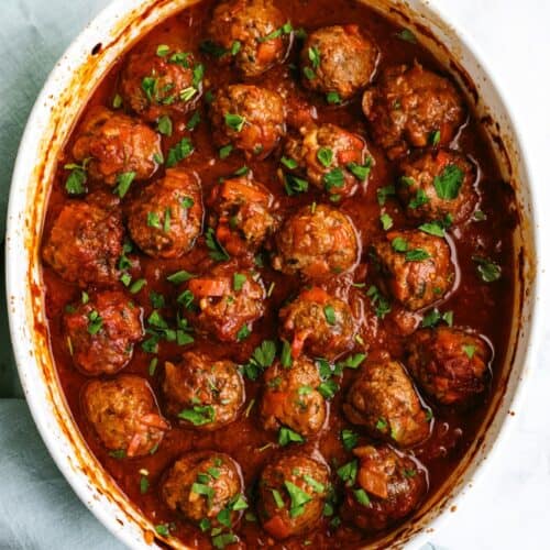 White casserole dish filled with meatballs in tomato sauce, topped with chopped parsley.