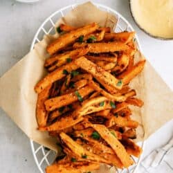 A basket of seasoned sweet potato fries garnished with herbs, placed on a piece of parchment paper, with two dipping sauces in bowls on the side.