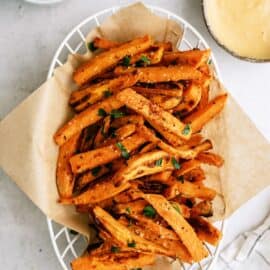 A basket of seasoned sweet potato fries garnished with herbs, placed on a piece of parchment paper, with two dipping sauces in bowls on the side.