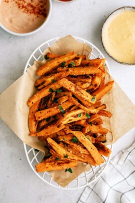 A basket of seasoned sweet potato fries garnished with herbs, placed on a piece of parchment paper, with two dipping sauces in bowls on the side.