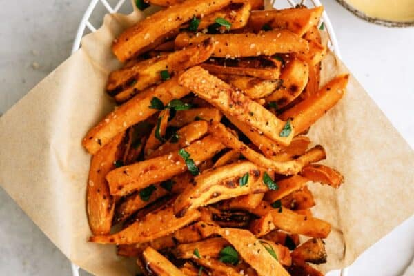 A basket of seasoned sweet potato fries garnished with herbs, placed on a piece of parchment paper, with two dipping sauces in bowls on the side.