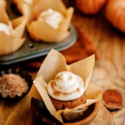 A pumpkin cupcake with cream cheese frosting, baked in a brown paper liner, is displayed on a wooden surface. In the background, a muffin tin with more cupcakes and small decorative pumpkins are visible.