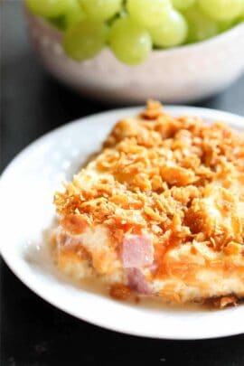 A serving of casserole with a crunchy topping on a white plate, with a bowl of green grapes in the background.