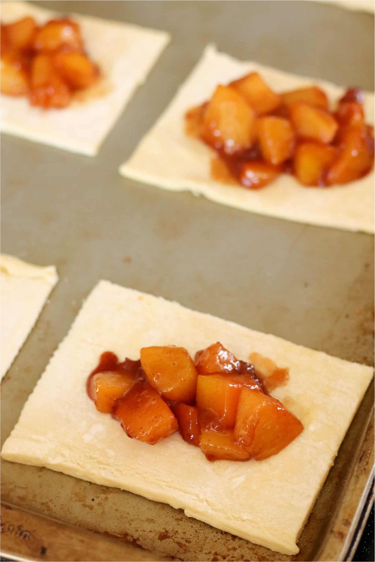 Small squares of puff pastry on a baking sheet with a scoop of peach pie filling in the middle.