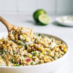 A white serving bowl filled with Mexican Street Corn Salad with a wooden spoon lifting a scoop out of the bowl. Lime sliced in half in the background.