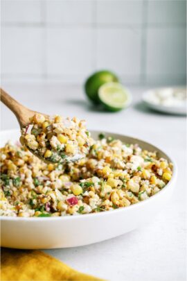 A white serving bowl filled with Mexican Street Corn Salad with a wooden spoon lifting a scoop out of the bowl. Lime sliced in half in the background.