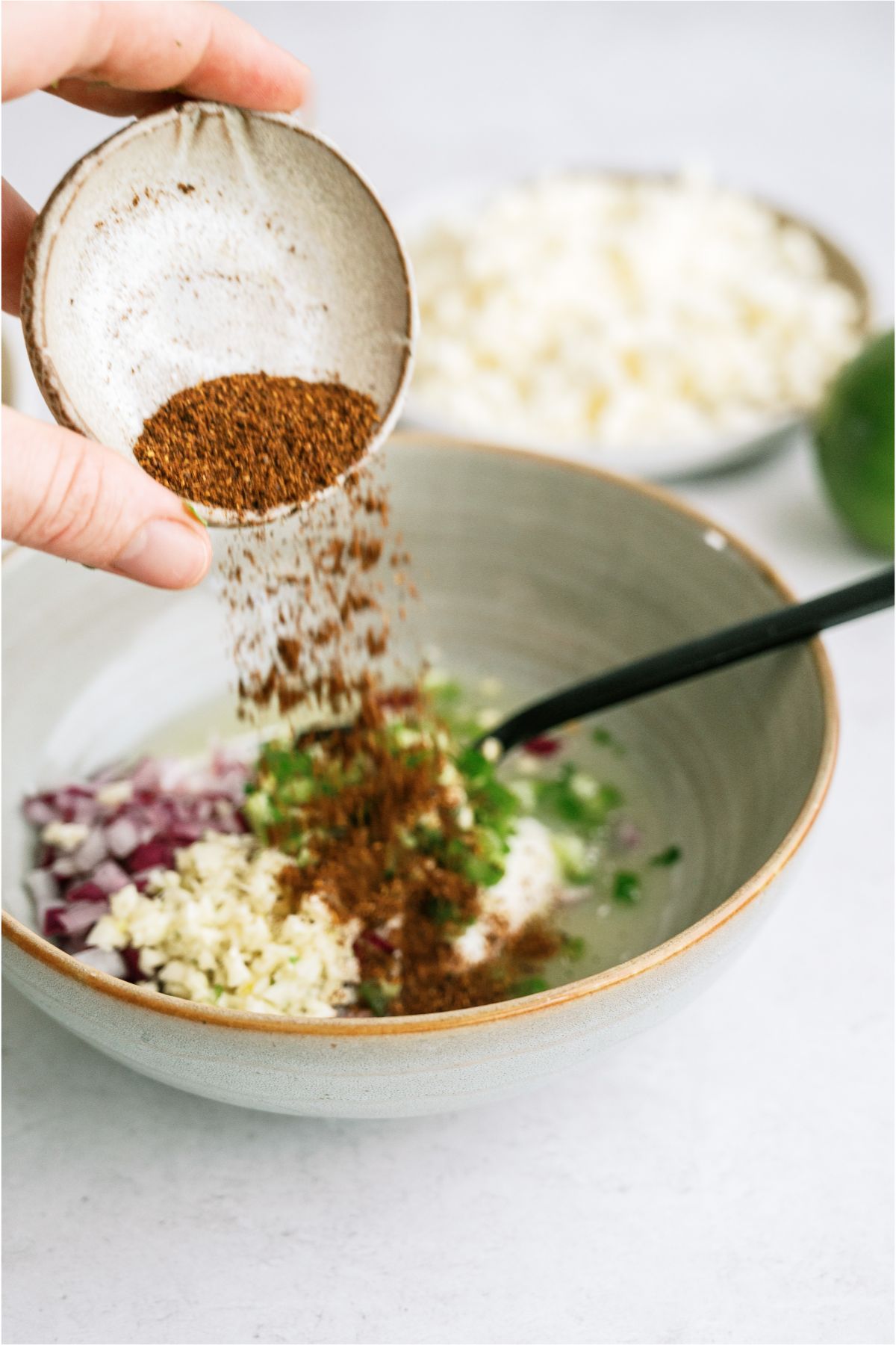 Pouring seasoning into a small bowl with other ingredients and a spoon inside the bowl.