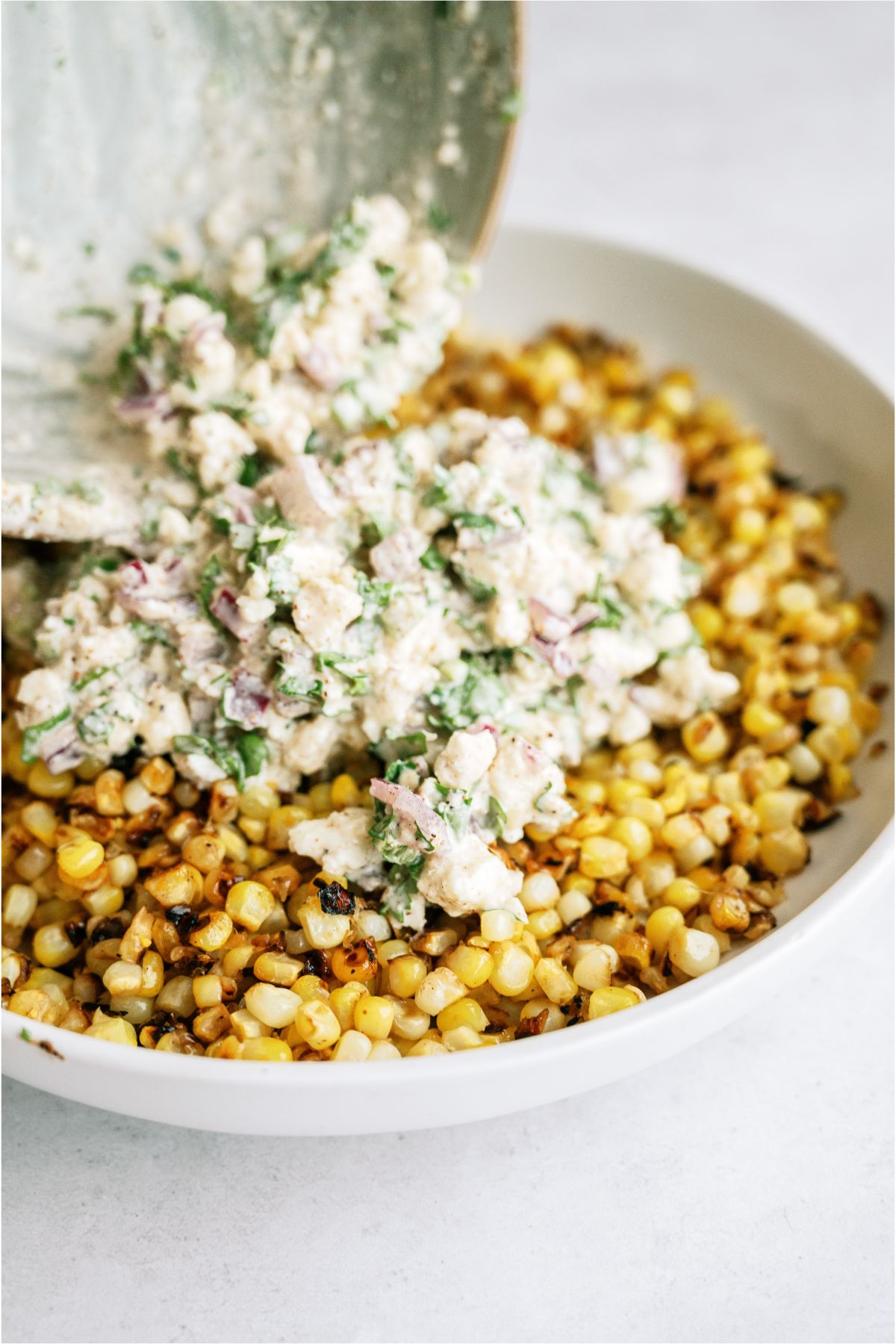 Pouring the cotija cheese mixture from the small bowl on top of the cooked corn in a large serving dish.