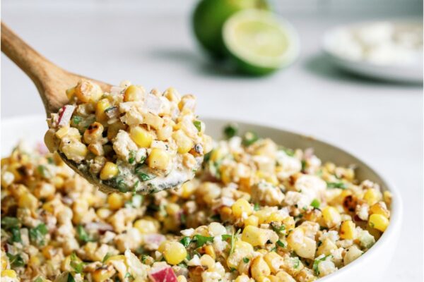 A white serving bowl filled with Mexican Street Corn Salad with a wooden spoon lifting a scoop out of the bowl. Lime sliced in half in the background.