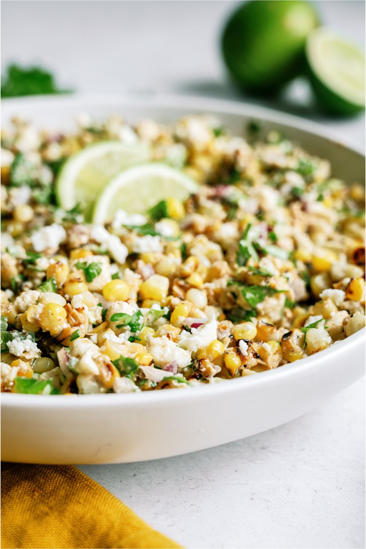 A large serving bowl filled with Mexican Street Corn Salad topped with 2 lime slices, with limes in the background.