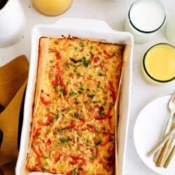 Baked casserole with red peppers and herbs in a white dish, surrounded by eggs, apples, orange juice, and a glass of milk on a table.