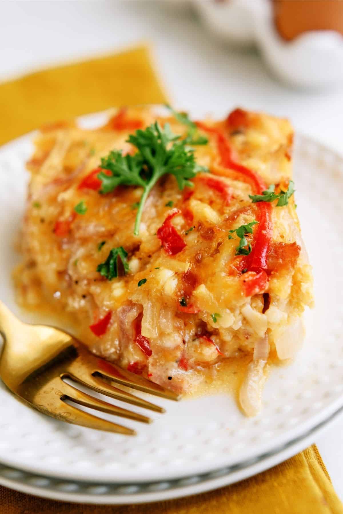 A slice of vegetable casserole topped with chopped parsley on a white plate, next to a fork.