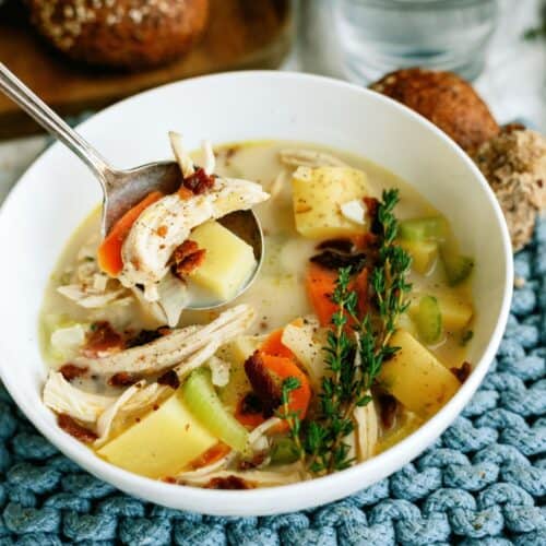 A white bowl filled with Creamy Chicken and Potato Soup with a spoon lifting out a bite. Rolls on a plate in the background.