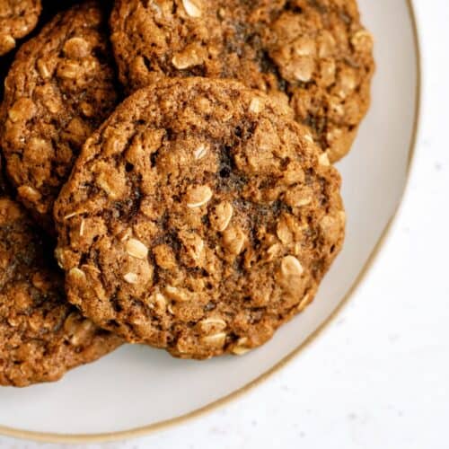 A close-up of a plate with several oatmeal cookies. The cookies have a coarse texture and visible oats. The plate is white with a gold rim and is placed on a white surface.