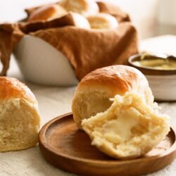 A roll on a plate cut in half with butter spread on the bottom half. A basket of rolls in the background.