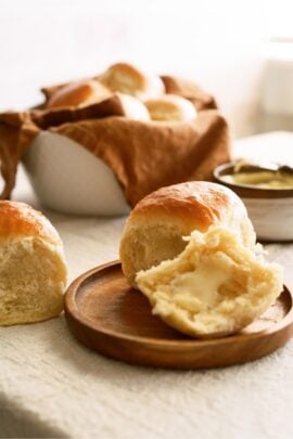 A roll on a plate cut in half with butter spread on the bottom half. A basket of rolls in the background.