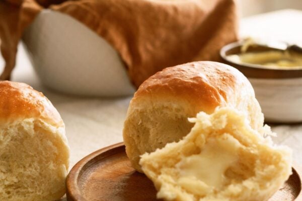 A roll on a plate cut in half with butter spread on the bottom half. A basket of rolls in the background.