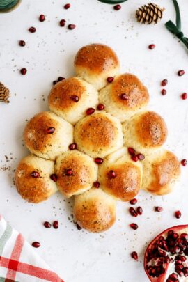 Bread rolls arranged in a Christmas tree shape, topped with herbs and pomegranate seeds, on a white surface with additional pomegranate seeds and pinecones scattered around.