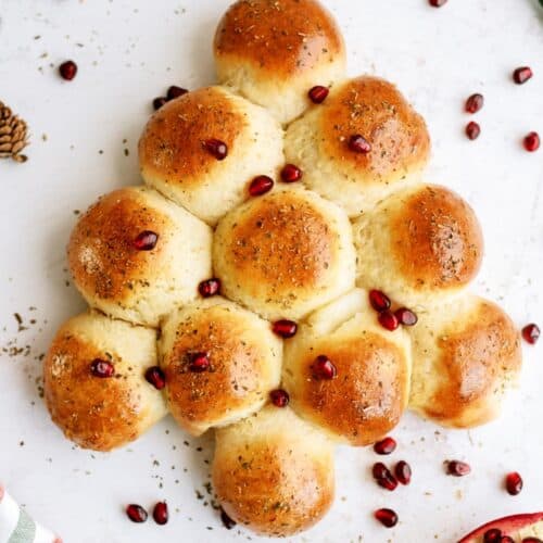 Bread rolls arranged in a Christmas tree shape, topped with herbs and pomegranate seeds, on a white surface with additional pomegranate seeds and pinecones scattered around.