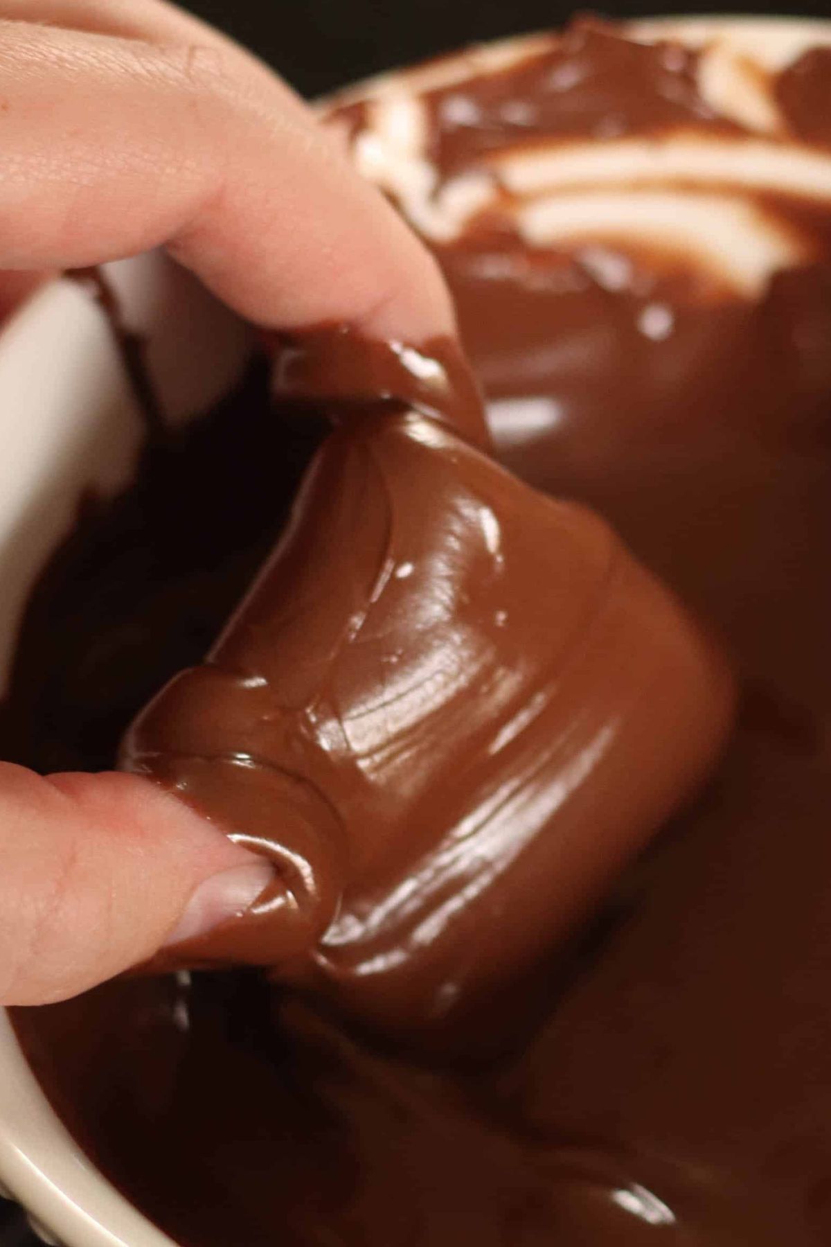 A hand dipping a caramel square in a bowl of melted milk chocolate.