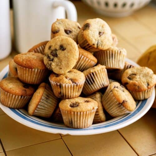 A plate filled with mini muffins, some with chocolate chips, sits on a tiled countertop. A mug, a bottle, and a bowl of bananas are in the background.